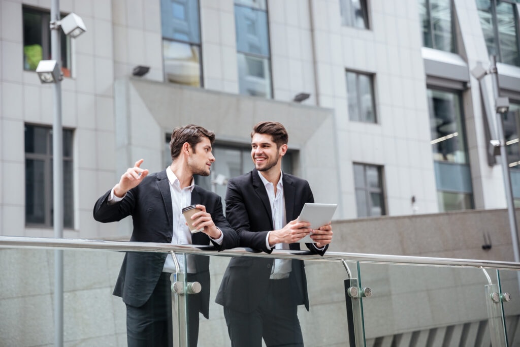 Two happy young businessmen with tablet standing and talking